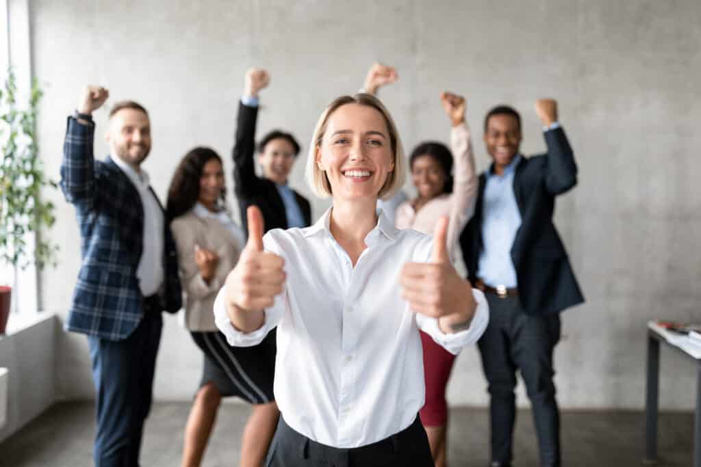 Businesswoman Gesturing Thumbs Up Standing With Joyful Employees In Modern Office.
